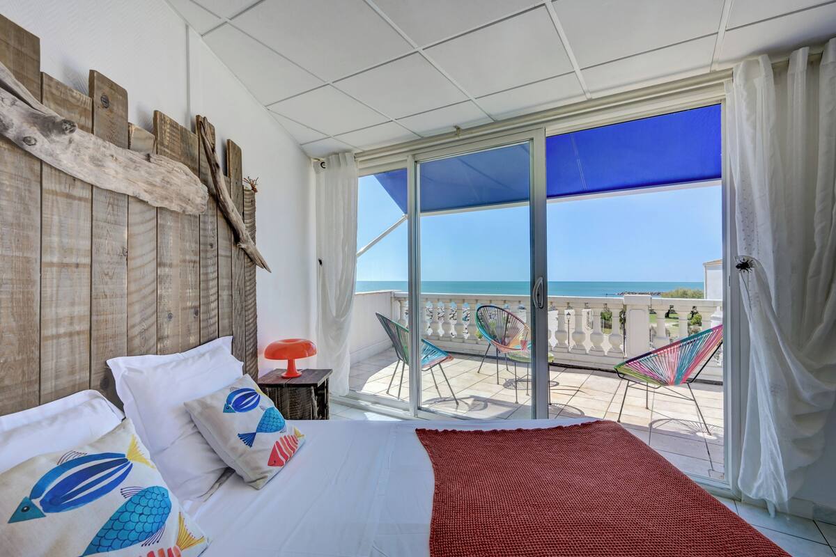 A bedroom with a large bed, a view of the sea through a sliding glass door, and a balcony with chairs and a table.