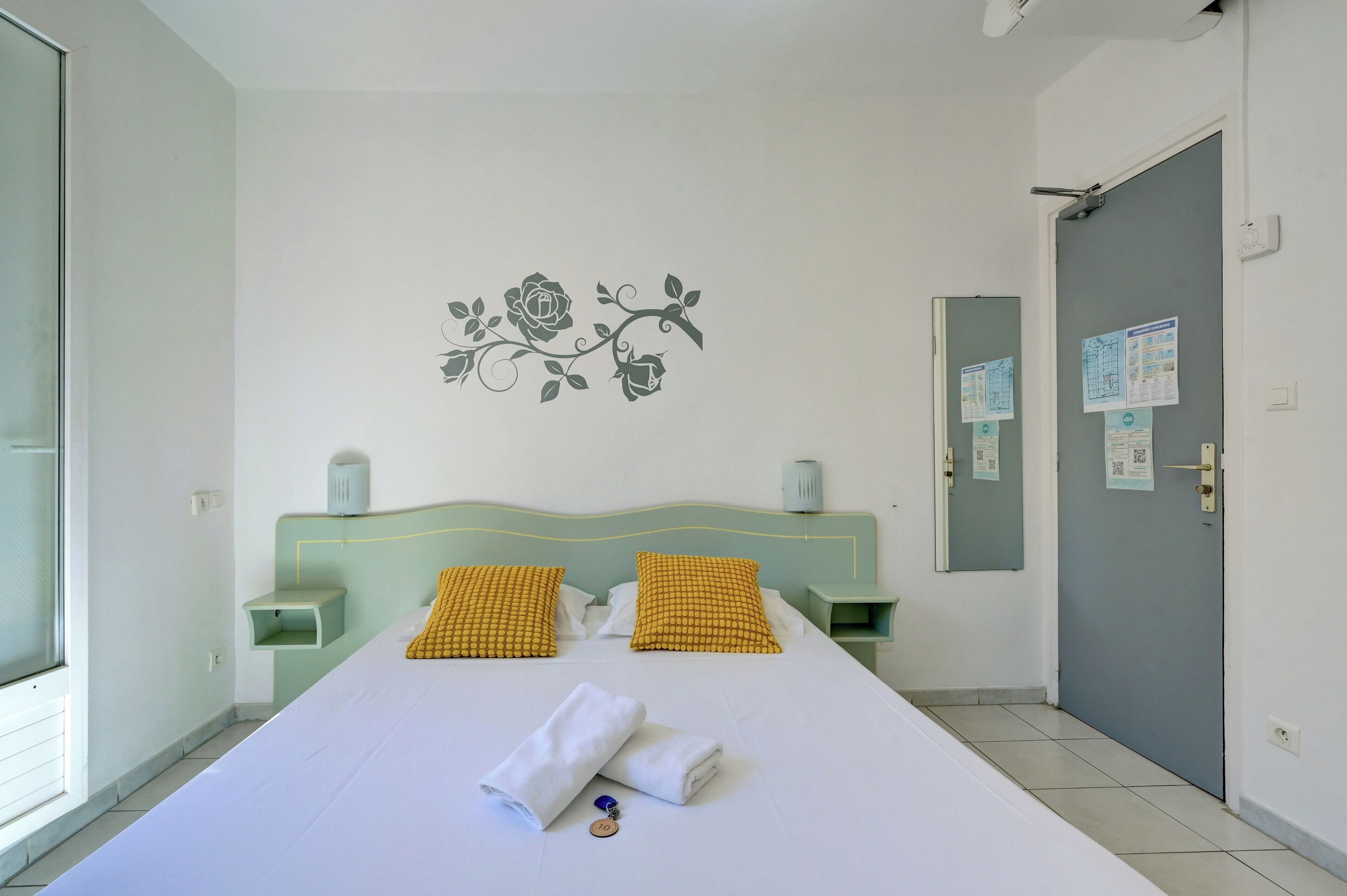 A hotel room with a bed, two pillows, a wall decoration, a bedside table, a nightstand, and a door with a mirror and papers.