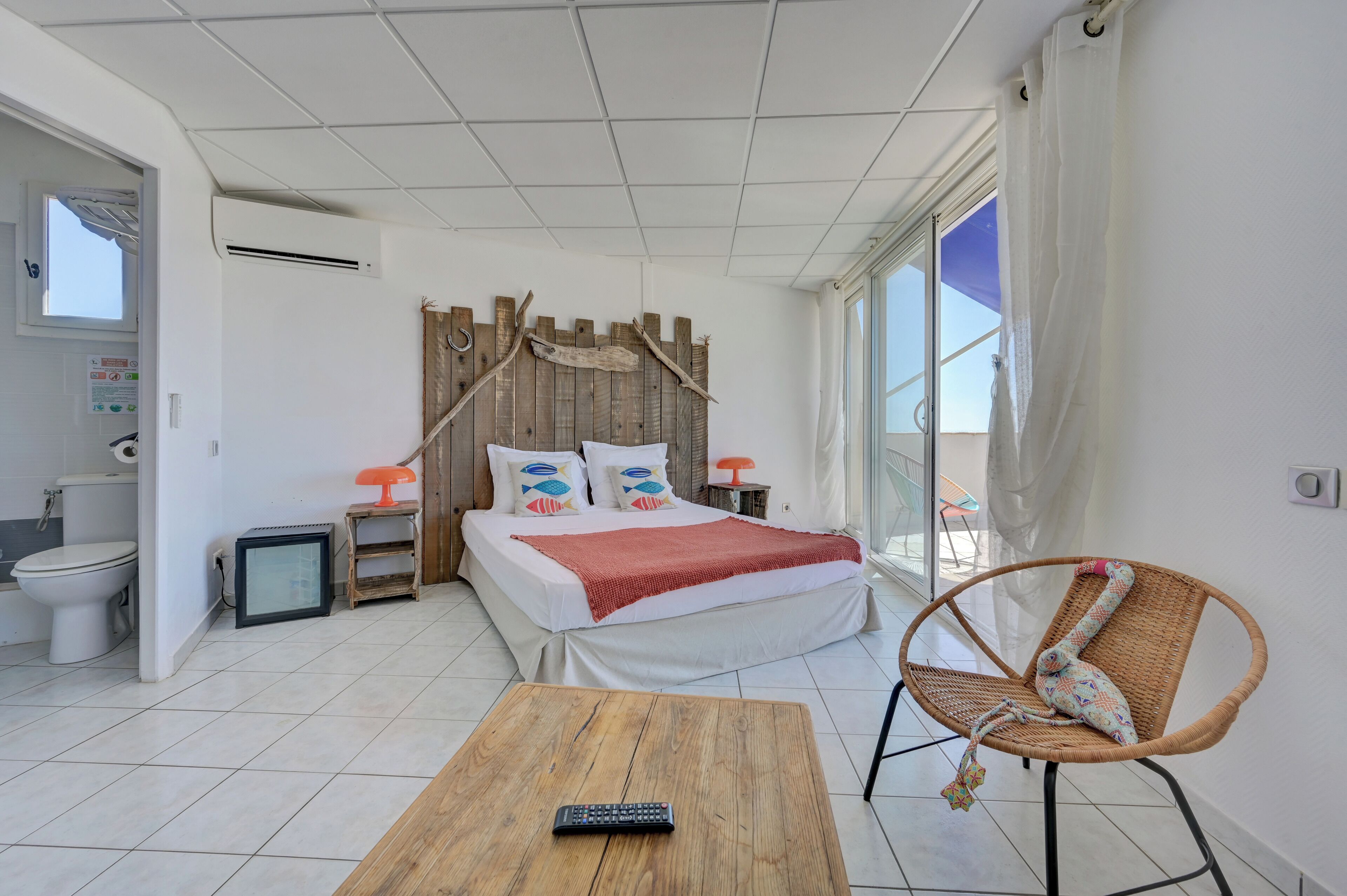A bedroom with a bed, a wicker chair, a small table with a remote control, a window with curtains, and a bathroom with a toilet and a mirror.