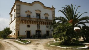 Un bâtiment historique à la façade symétrique, doté d’une entrée centrale et entouré d’un jardin planté de palmiers.