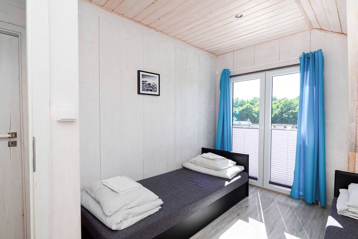 A room with two beds, a wooden ceiling, a window with blue curtains, and a framed picture on the wall.