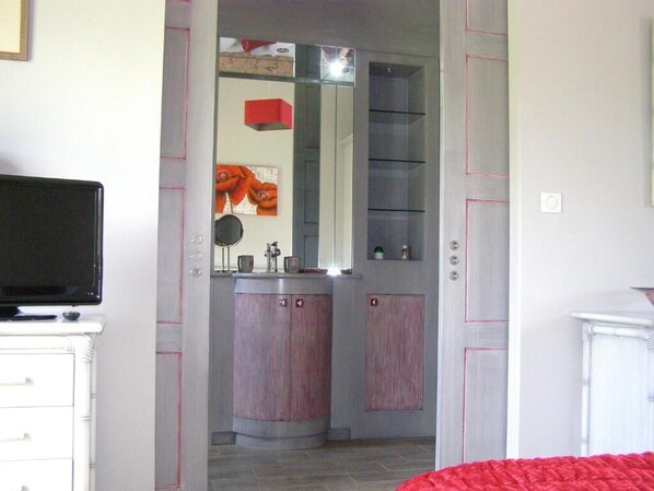 Una habitación con cama, televisión, espejo y baño con lavamanos y armarios.