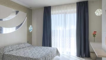 A hotel room with a bed, a large window with curtains, a flat-screen TV on the wall, and a small table with a vase of flowers.