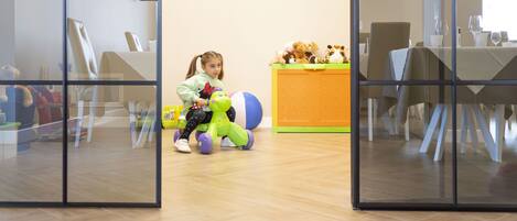 Un niño jugando con juguetes en una habitación con un ventanal grande, una mesa con sillas y un mostrador lleno de peluches.