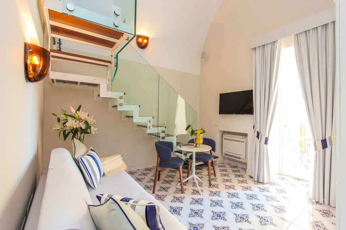 A modern hotel room with a glass staircase, a sofa with patterned cushions, a dining table with chairs, and a flat-screen TV mounted on the wall.