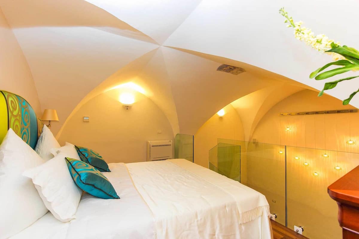 A bedroom with a curved ceiling, a bed with white bedding and teal pillows, a bedside lamp, and a wall-mounted air conditioner.