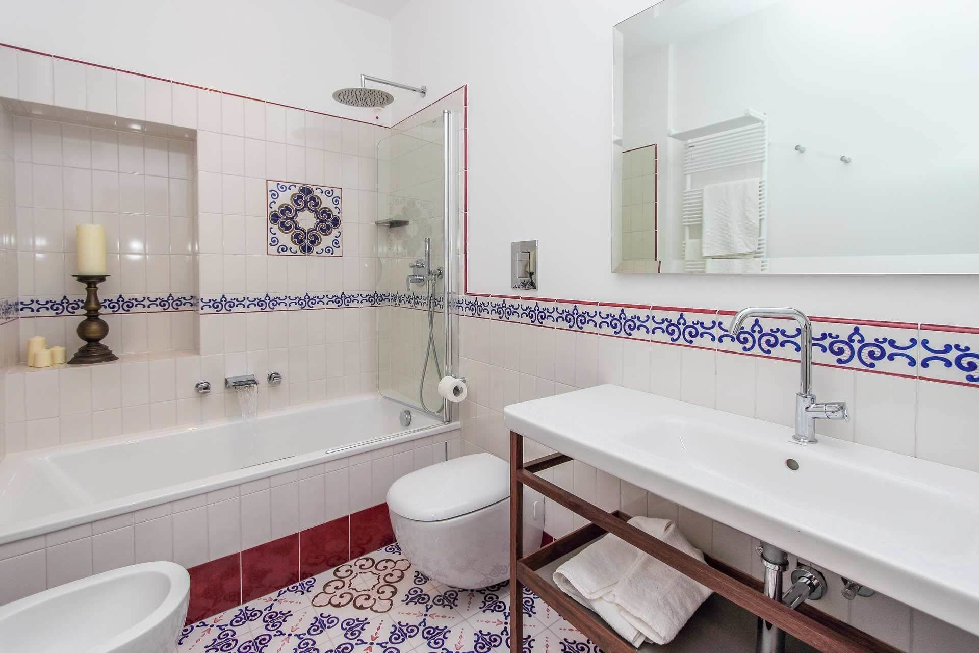 A bathroom with a bathtub, toilet, and sink, featuring white tiles with blue and red decorative borders.