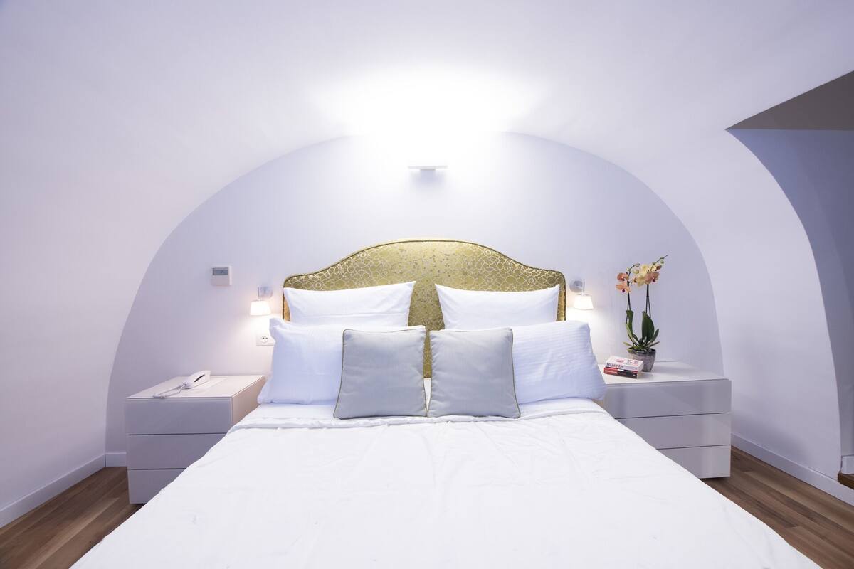A modern bedroom with a large bed, white bedding, a gold-patterned headboard, and two bedside tables with lamps.