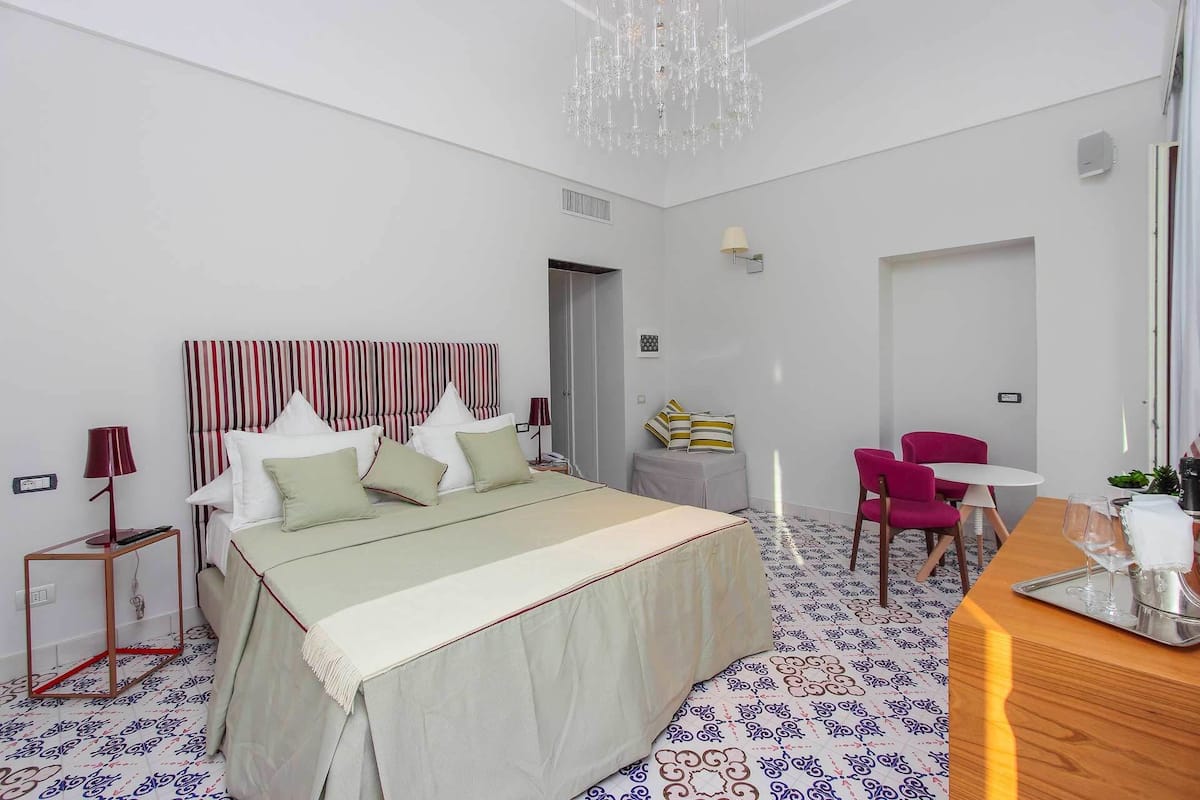 A hotel room with a bed, bedside tables, a desk, chairs, a chandelier, and a patterned floor.