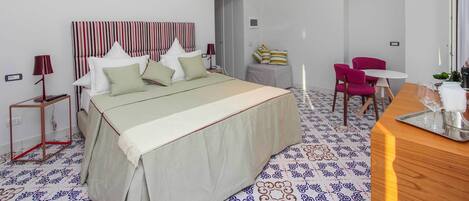 A hotel room with a bed, bedside tables, a desk, chairs, a chandelier, and a patterned floor.