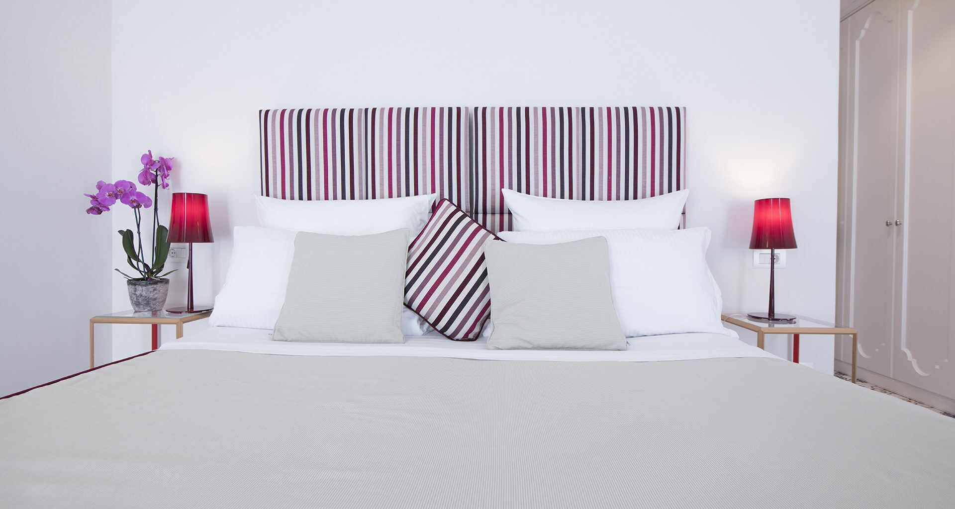 Una cama bien hecha con ropa blanca y un cabecero a rayas, dos mesitas de noche con lámparas y una orquídea en maceta a la izquierda.