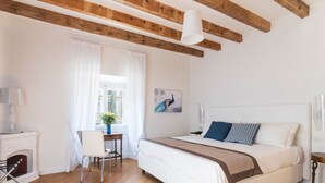 A bedroom with a large bed, a desk, a chair, a fireplace, and wooden beams on the ceiling.