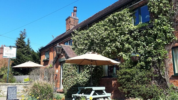 Un bâtiment en briques dont les murs sont recouverts de lierre, avec des tables à l’extérieur, protégées par des parasols, et une enseigne indiquant qu’il s’agit d’une maison d’hôtes et d’un salon de thé.