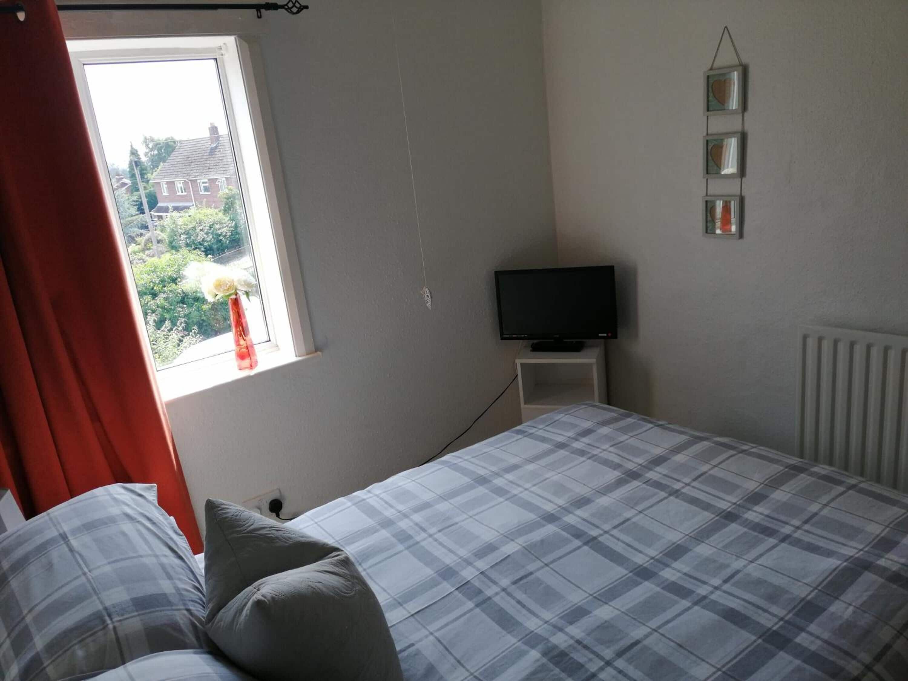 A bedroom with a bed, a television on a small table, a window with curtains, and a radiator.