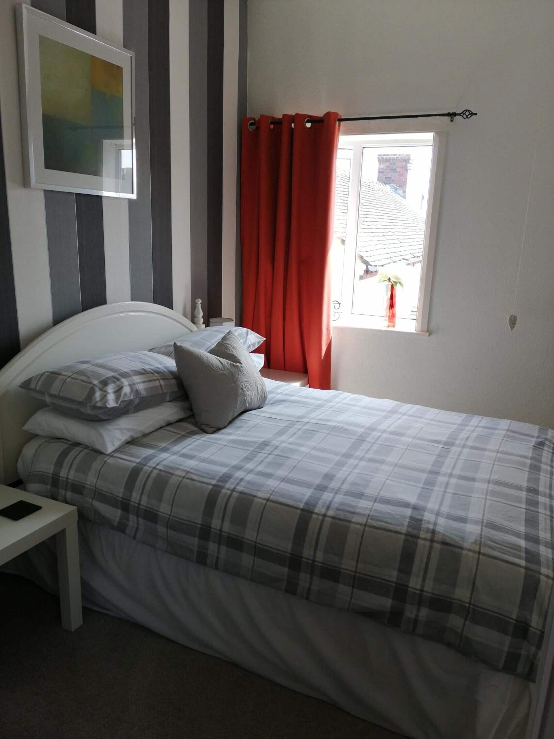 Una camera da letto con un letto, cuscini, un comodino, una finestra con tende rosse e un quadro appeso al muro.