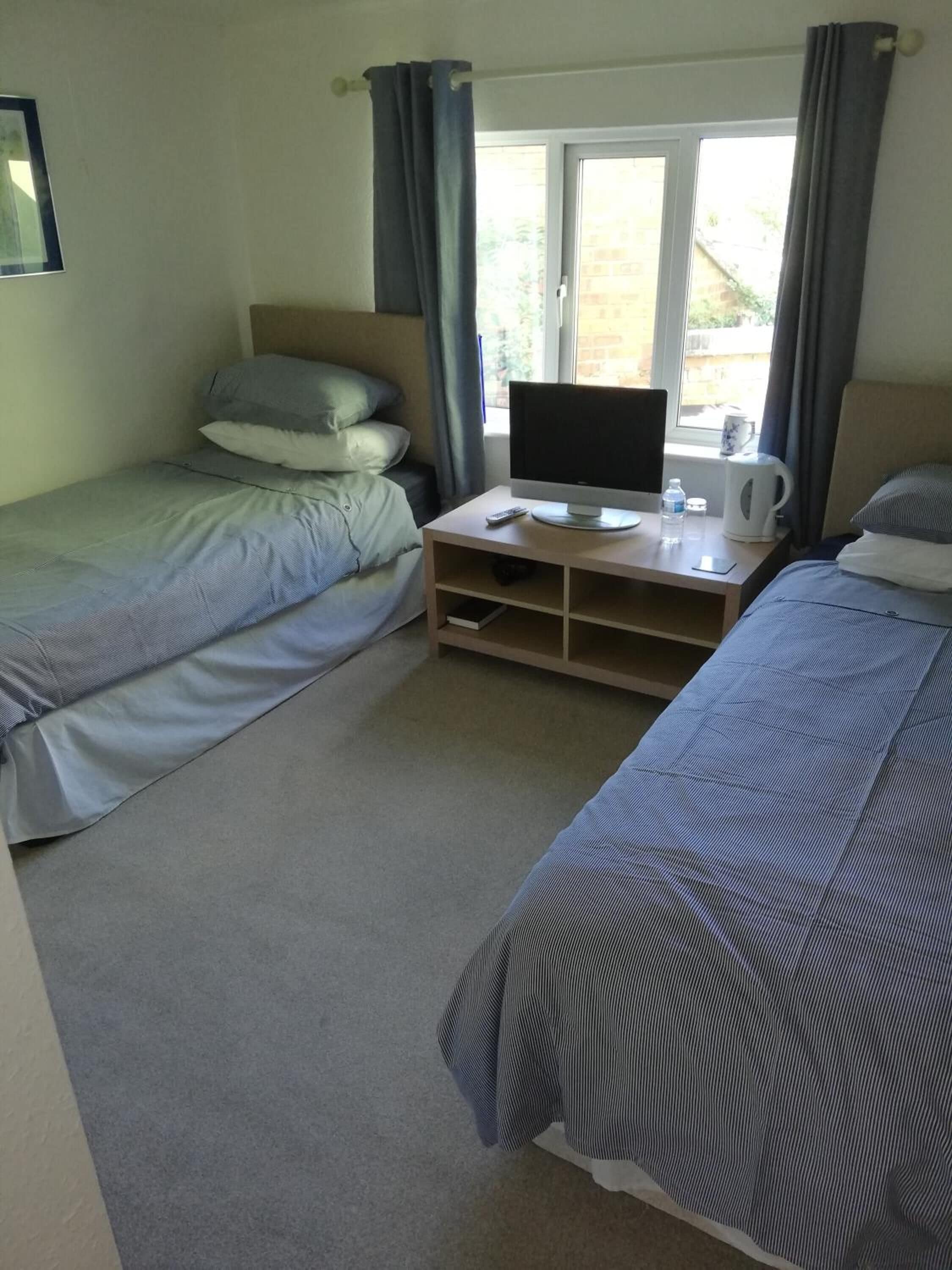 A room with two single beds, a TV on a wooden stand, a kettle, and a window with curtains.
