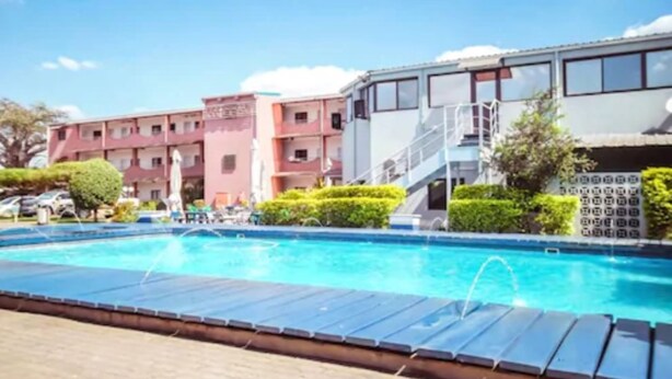 A swimming pool with a water fountain, surrounded by lounge chairs and a building with multiple floors and balconies. - Hotel Moatize (Moatize)