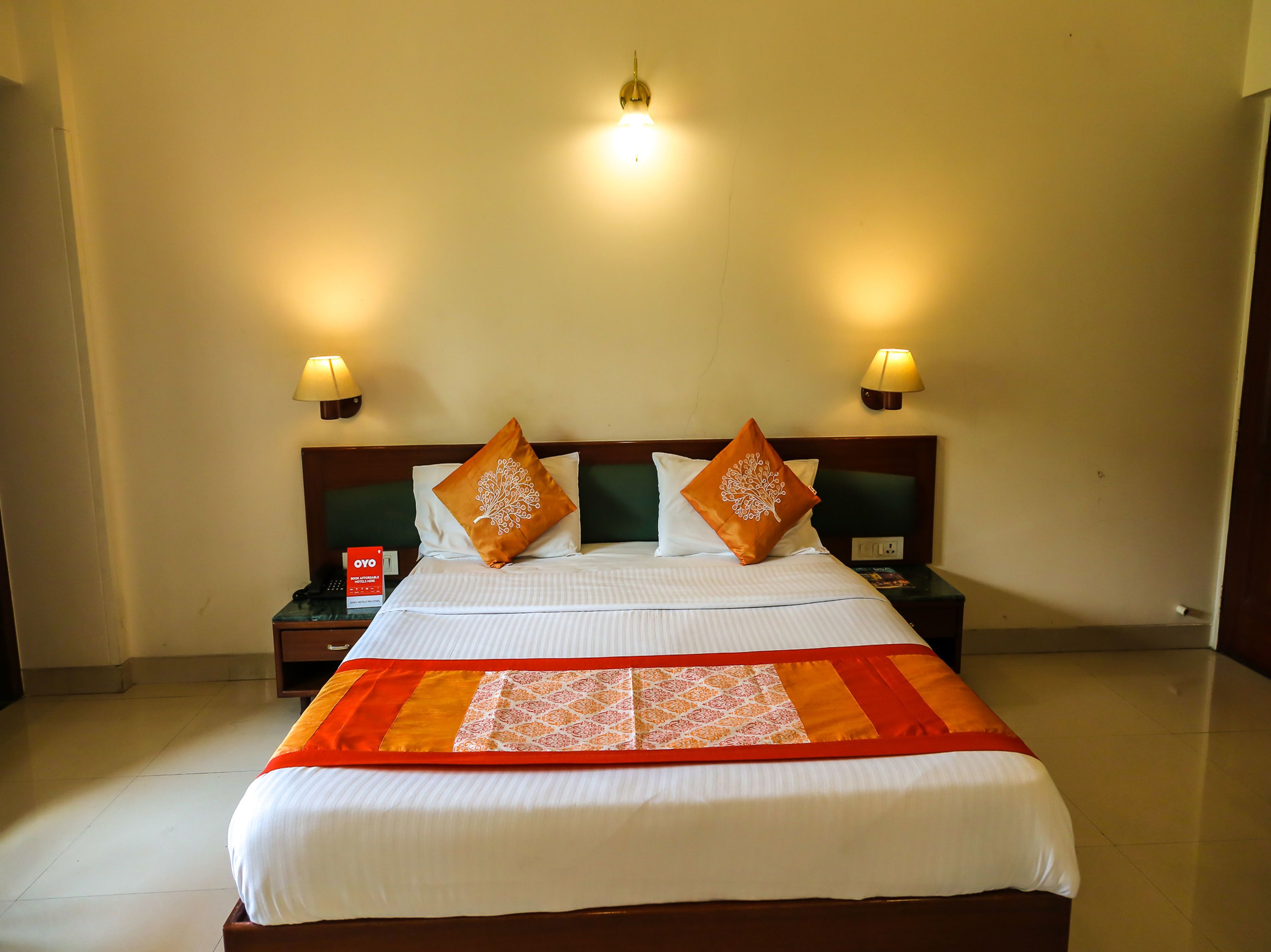 A neatly made bed with two decorative pillows, a bedside table with a lamp, and a wall-mounted light fixture.