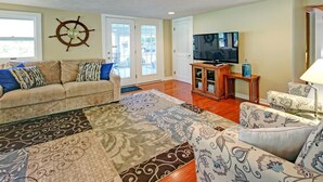 Una sala de estar con un sofá color crema, sillones estampados, un mueble de entretenimiento de madera con un televisor y un adorno de timón de barco en la pared.