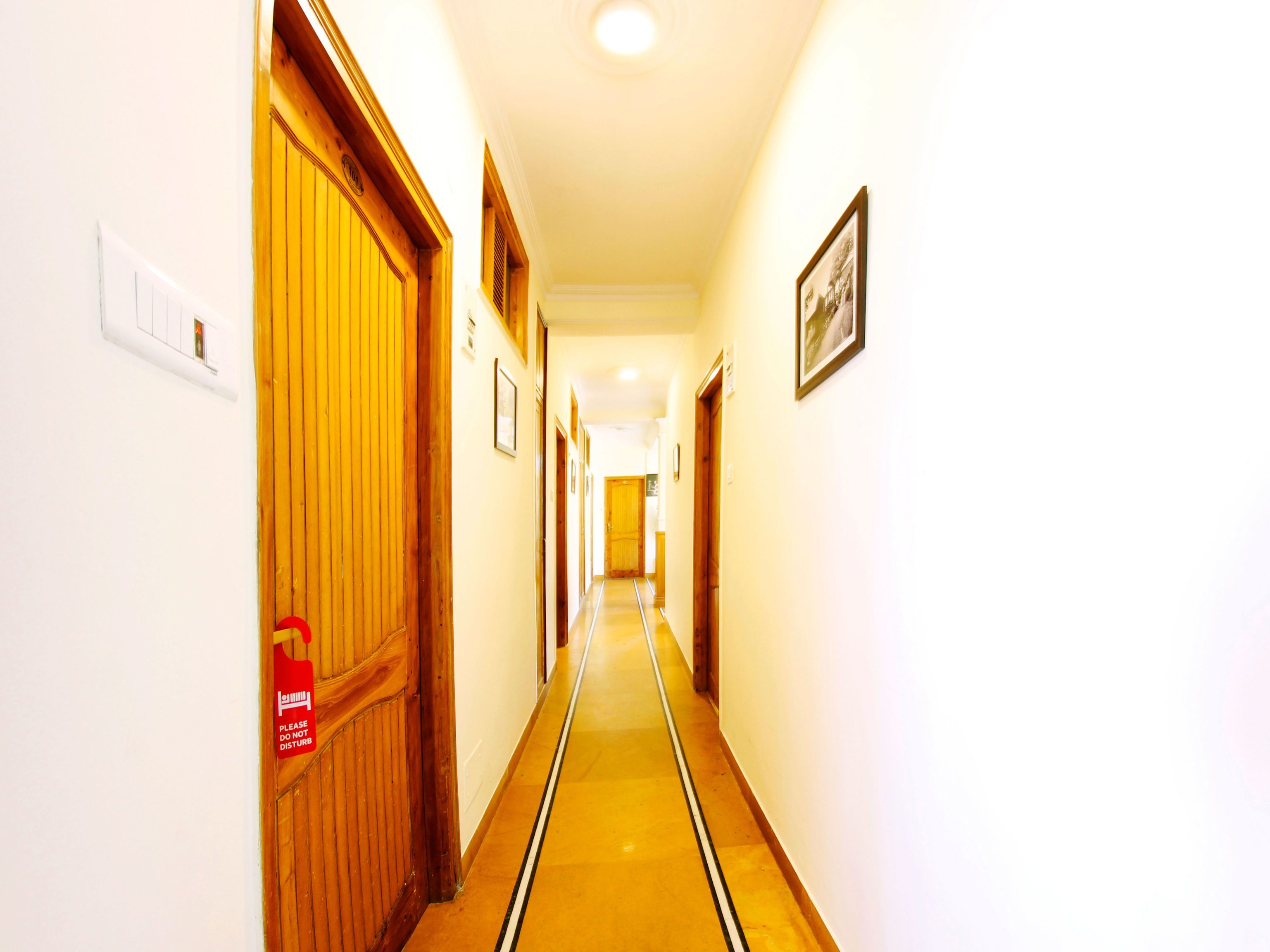Un couloir d’hôtel avec des portes en bois, un détecteur d’incendie et un tableau encadré accroché au mur.