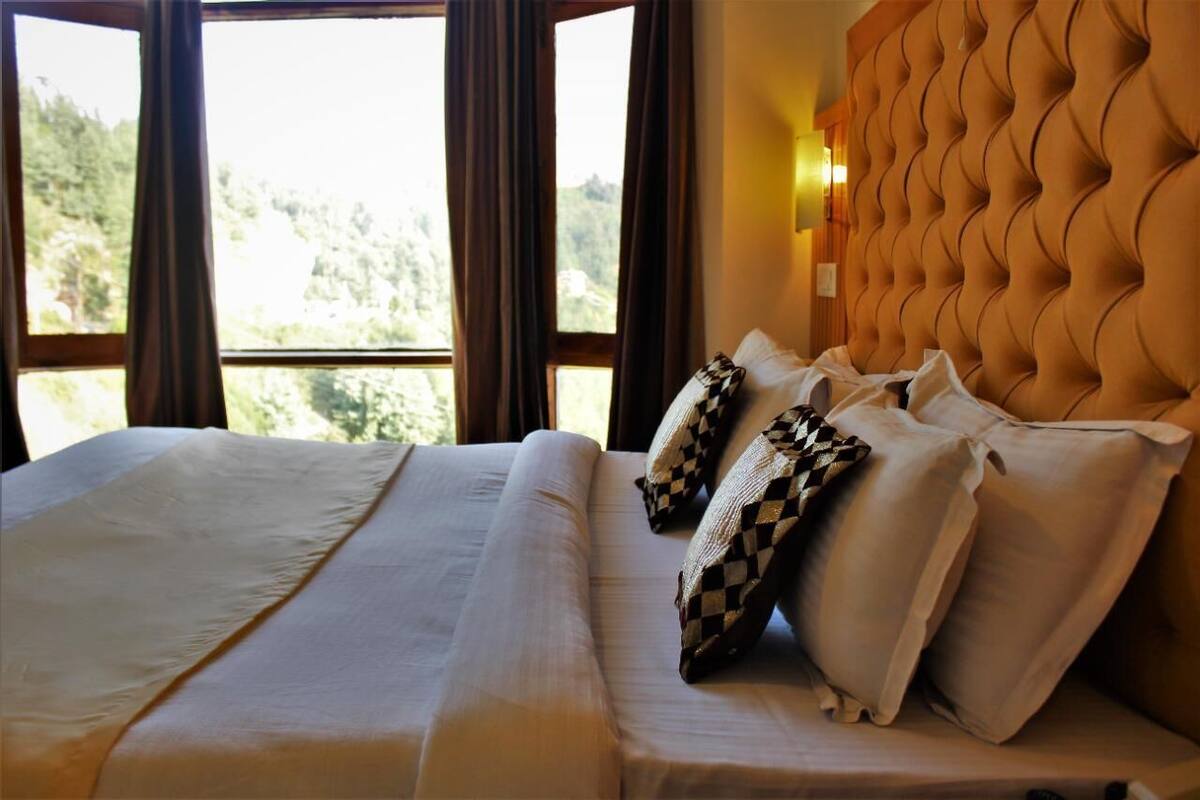 Ein Hotelzimmer mit einem großen Bett, einem gebuschten Kopfteil und Blick auf Bäume durch das Fenster.