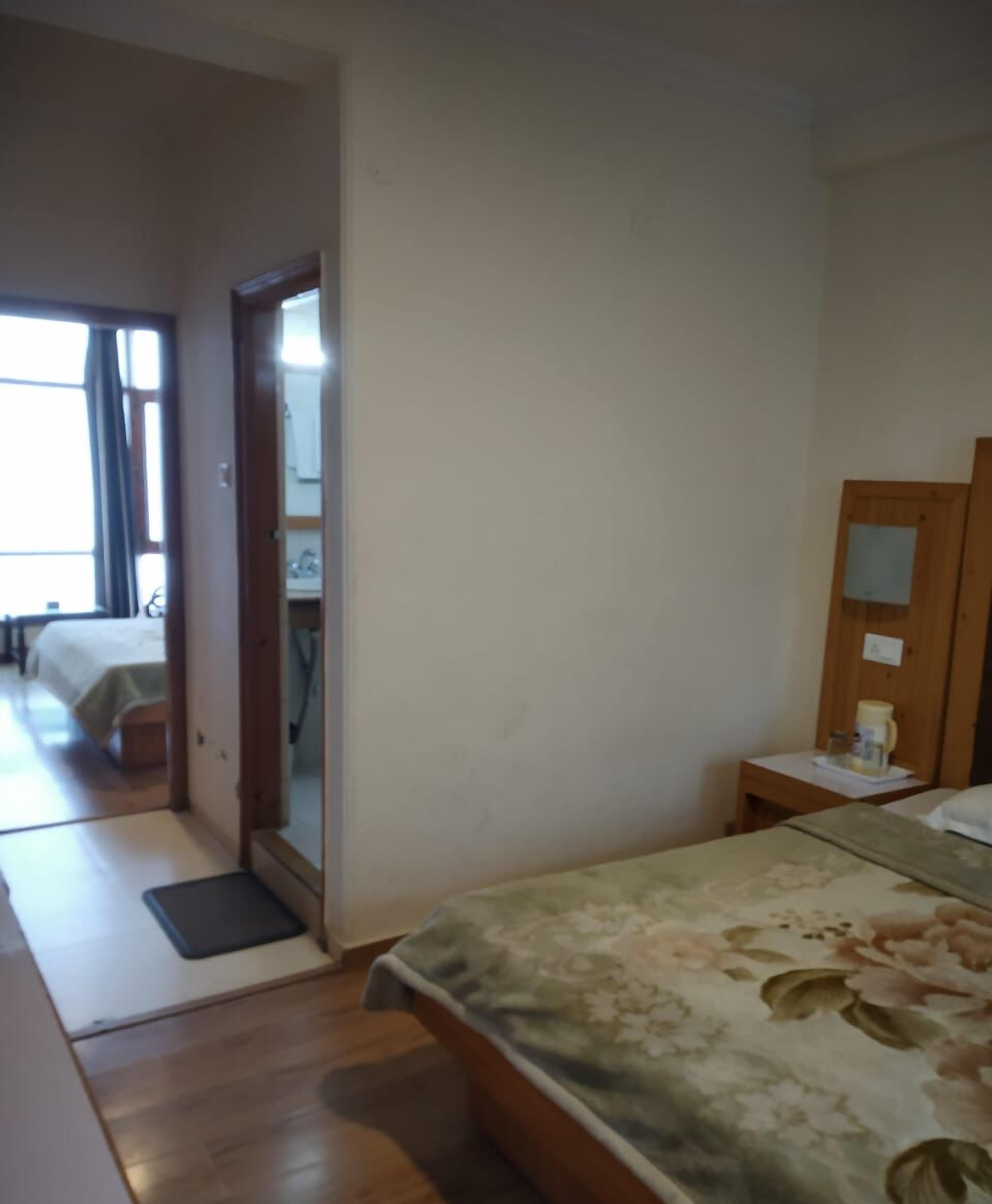 Habitación de hotel con cama, mesita de noche con lámpara, espejo y vista a otra habitación con cama y cortinas.