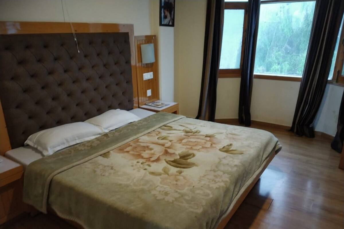 Ein Schlafzimmer mit einem gepolsterten Kopfende, einer blumigen Bettdecke, einem Nachttisch und einem Fenster mit Vorhängen.