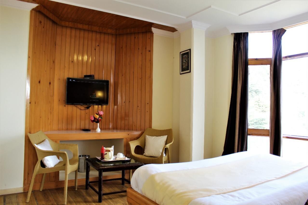 Een hotelkamer met een wand van houten panelen, een bed, twee stoelen, een bureau met een televisie en een raam met gordijnen.