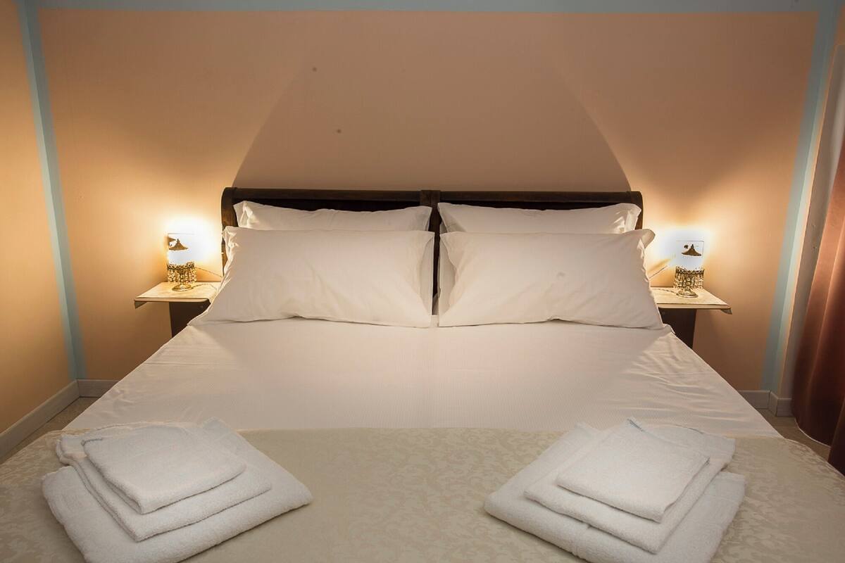 Un lit bien fait, avec du linge de lit blanc et des oreillers, deux tables de chevet avec des lampes, et des serviettes pliées posées sur le lit.
