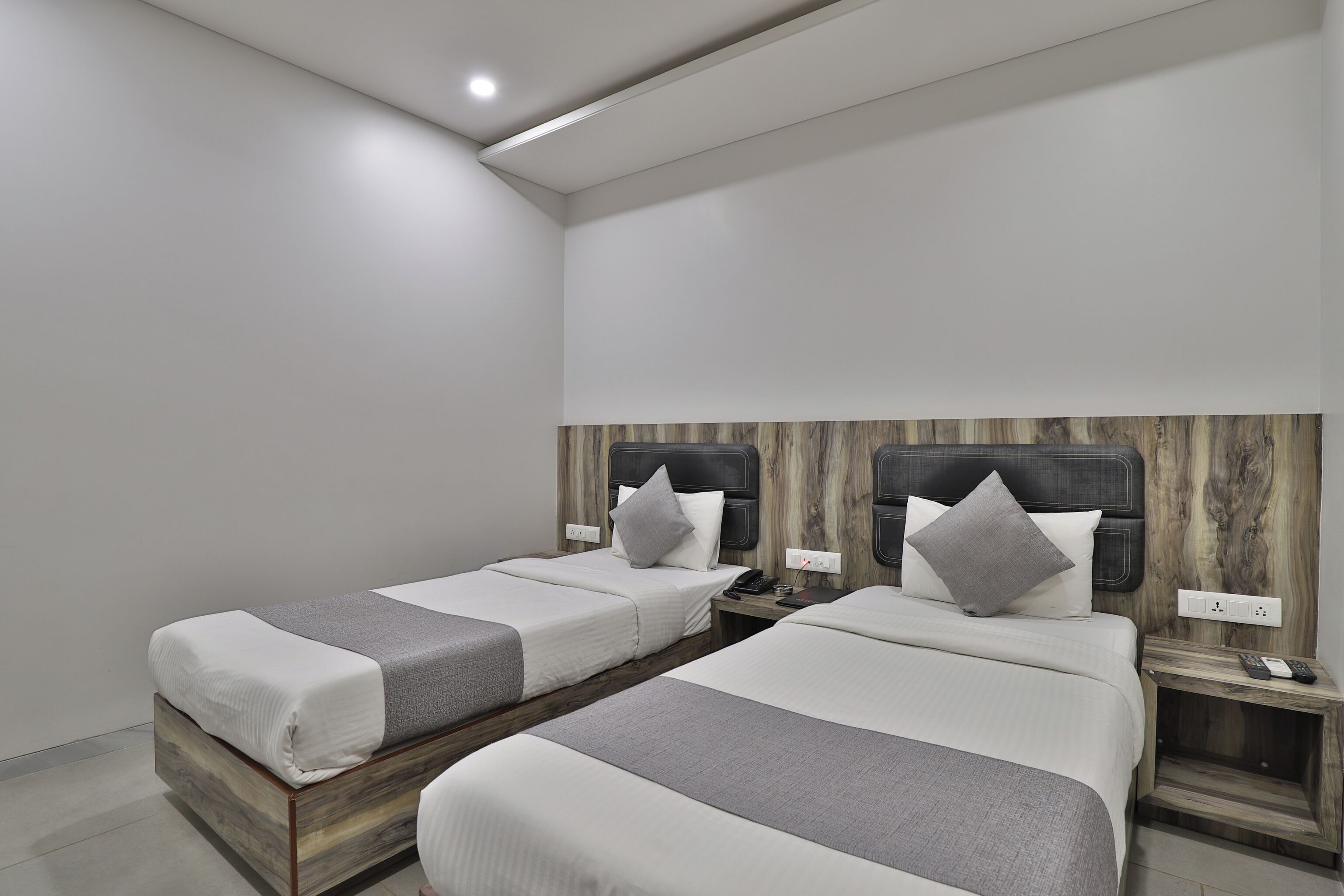 Habitación de hotel con dos camas, cada una con cabecero de madera, ropa de cama blanca y una manta gris. Hay mesitas de noche con lámparas y un teléfono.