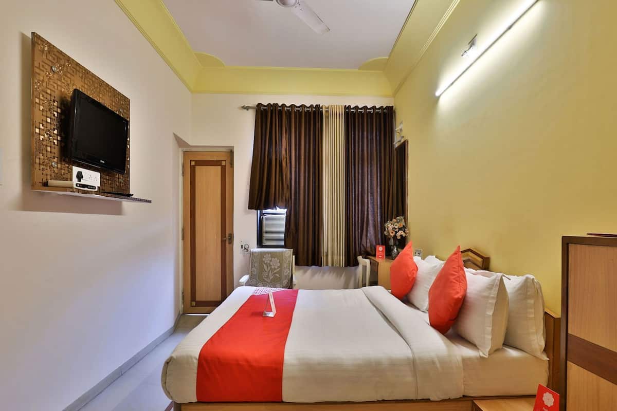 Une chambre d’hôtel avec un lit, un téléviseur fixé au mur, une fenêtre avec des rideaux et une table de chevet ornée d’un vase de fleurs.