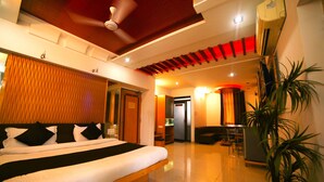 A hotel room with a large bed, a ceiling fan, a red accent wall, a plant, and a kitchenette. - Hotel Vrundavan Residency (Vadodara)