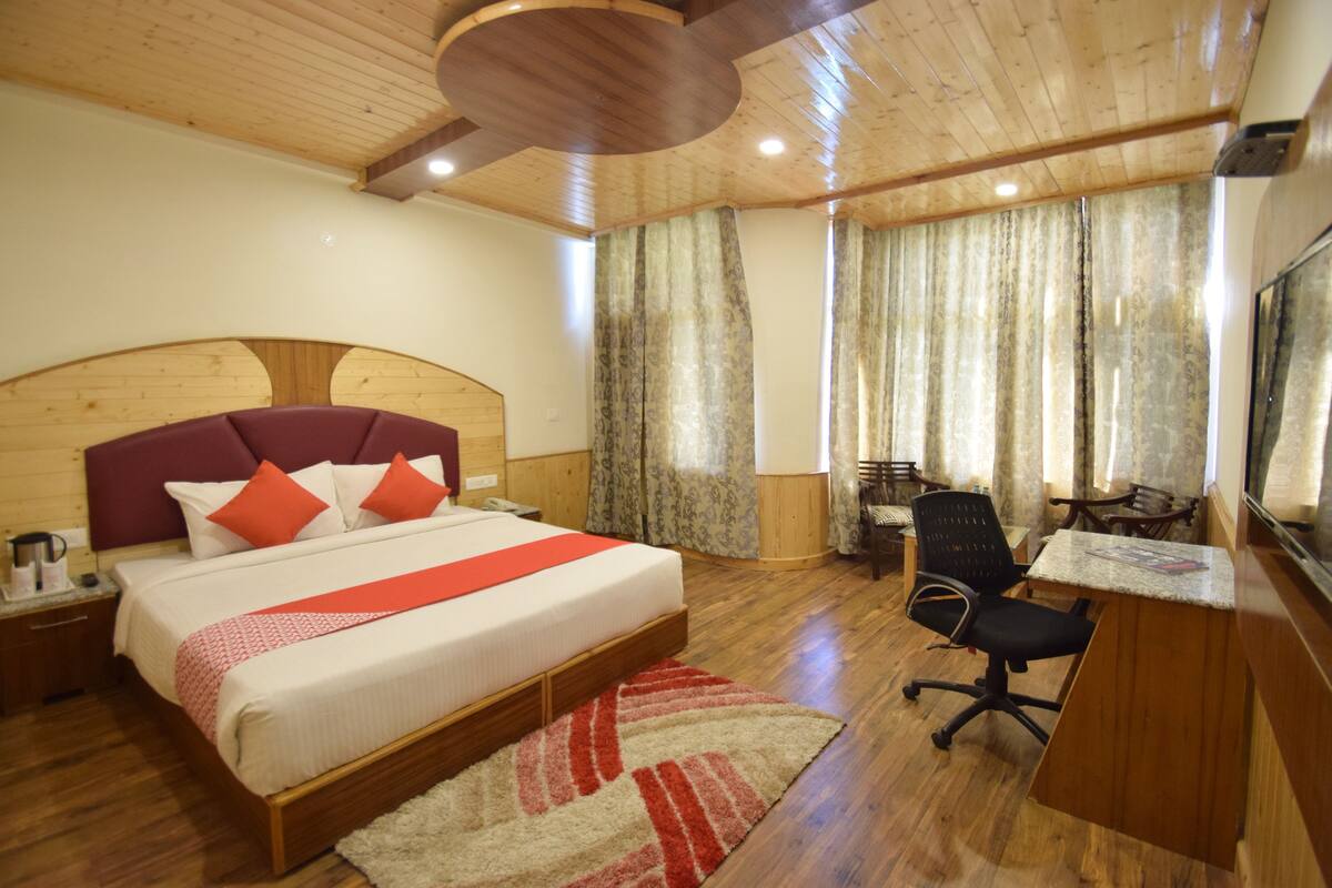 Un dormitorio con cabecera de madera, cama con ropa de cama roja y blanca, escritorio con silla y una ventana grande con cortinas.