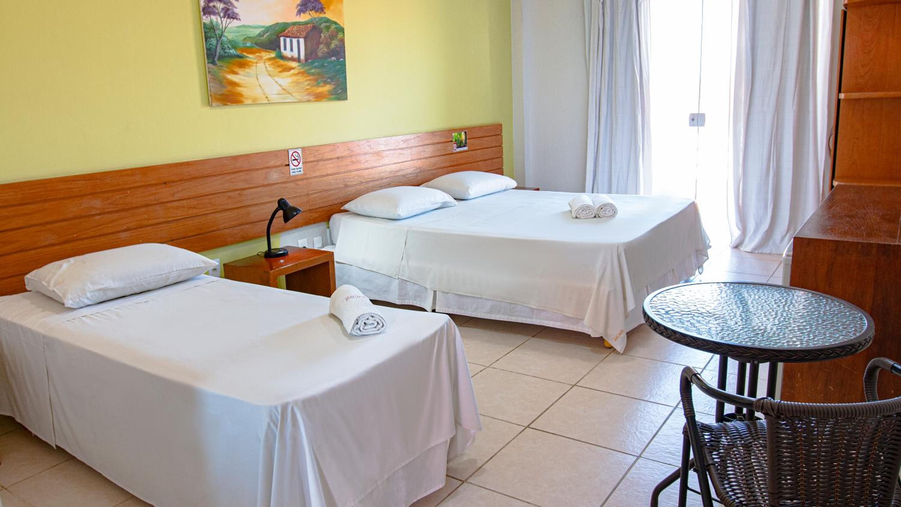 A hotel room with two single beds, a small table, a chair, and a painting on the wall.