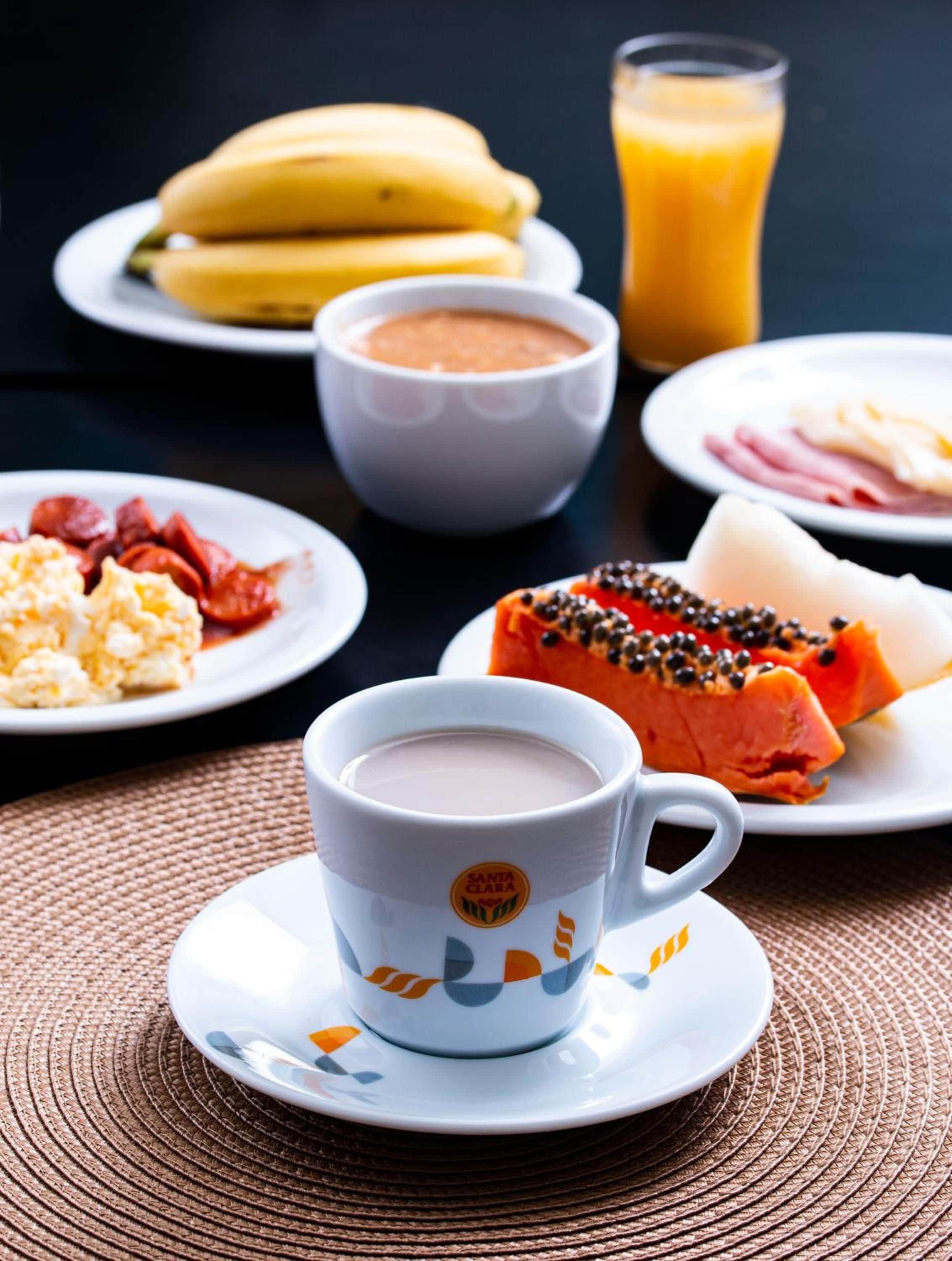 Une table mise pour le petit-déjeuner avec une tasse de café, un verre de jus d’orange et des assiettes contenant des œufs, du jambon et un assortiment de fruits.