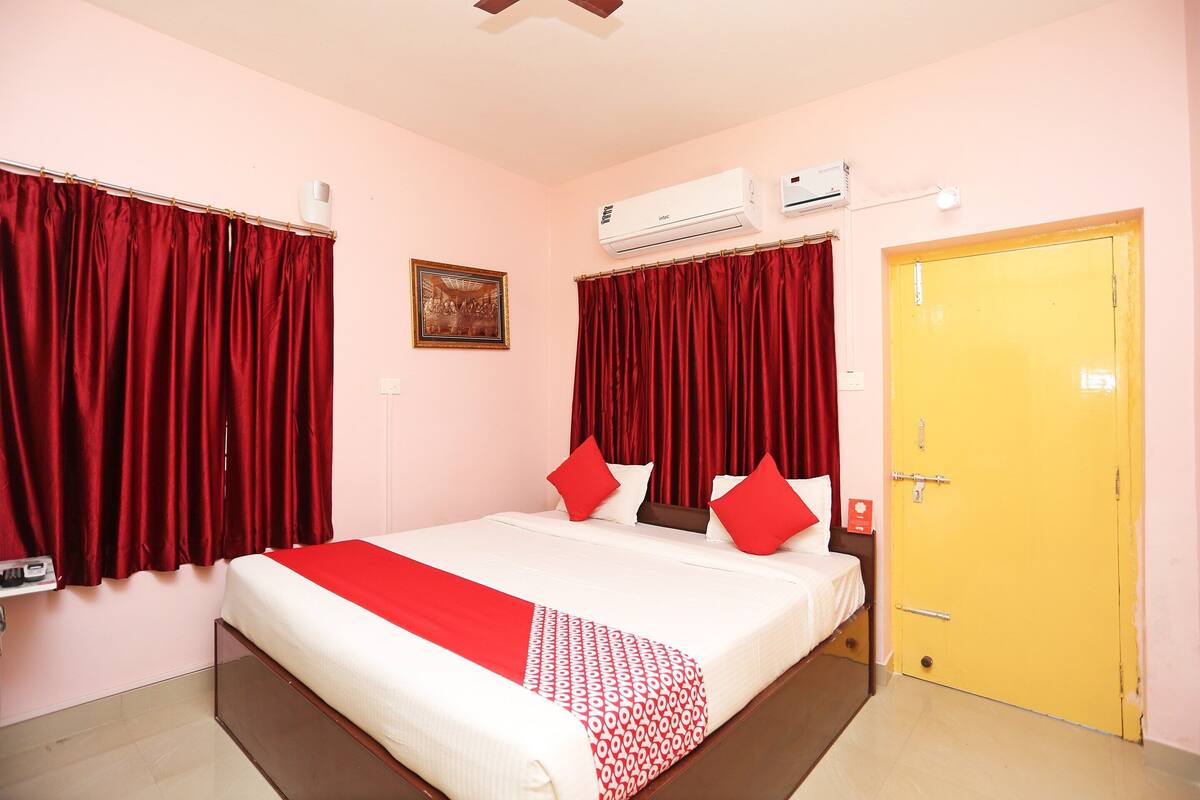 A hotel room with a bed, red curtains, an air conditioning unit, a yellow door, and a framed picture on the wall.