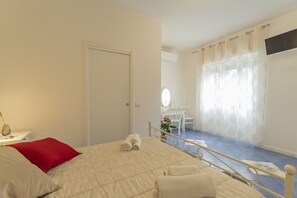 Un dormitorio con una cama, una mesita de noche, un almohadón rojo y una ventana con cortinas.
