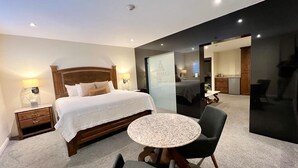A modern hotel room with a large bed, a round table, and a chair. There is a nightstand with a lamp and a mirror. The room has a carpeted floor and a dark accent wall. - Hotel Caesar's (Tijuana)