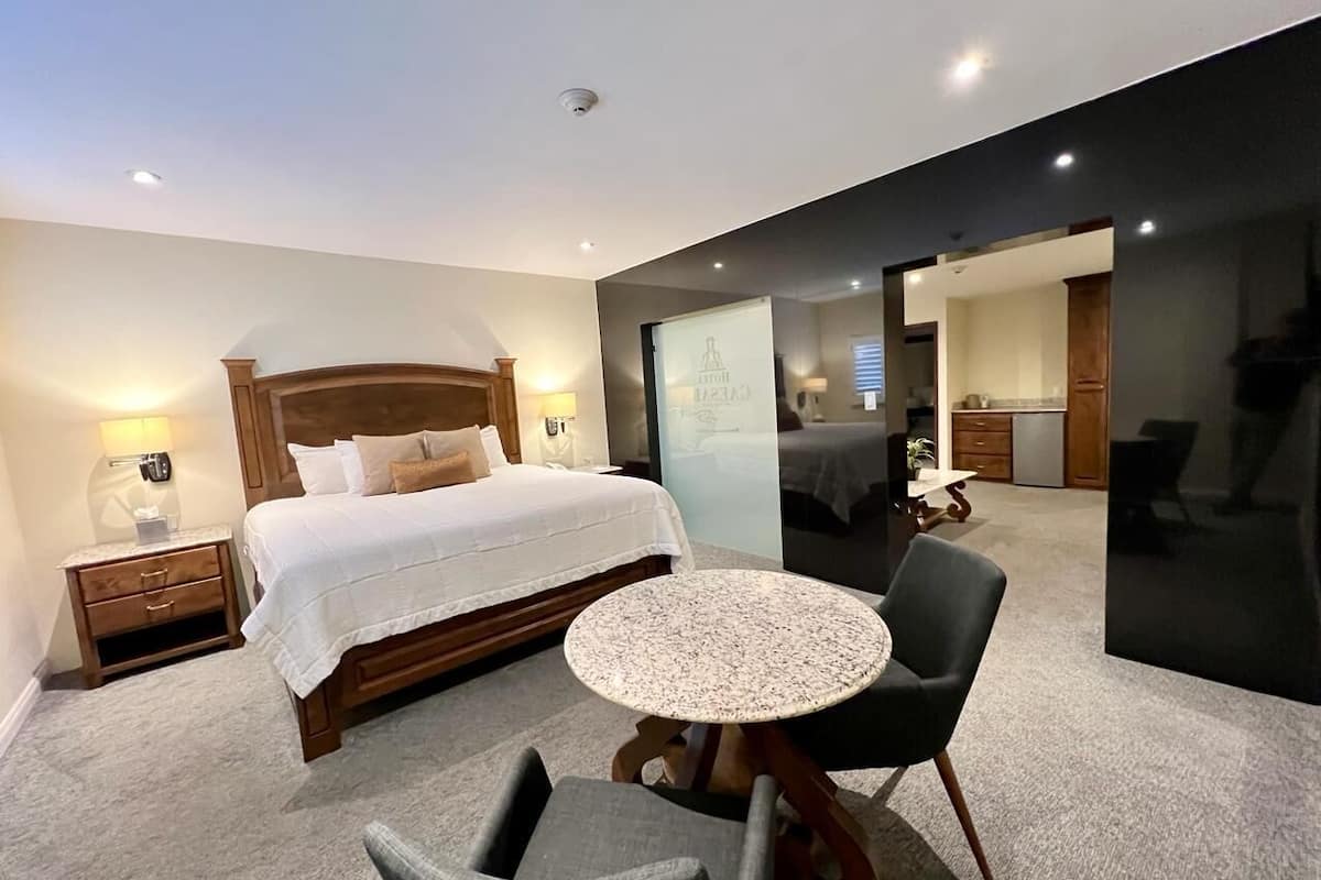 A modern hotel room with a large bed, a round table, and a chair. There is a nightstand with a lamp and a mirror. The room has a carpeted floor and a dark accent wall.