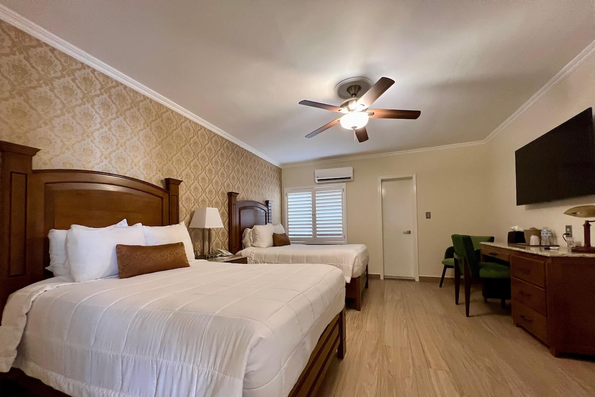 A hotel room with two beds, a desk, a chair, a ceiling fan, and a TV mounted on the wall.