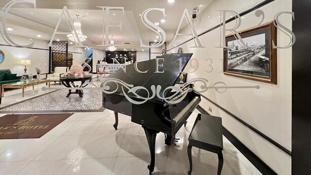 A grand piano in a well-lit lobby with a modern chandelier, a seating area with a green sofa, and a framed picture on the wall. - Hotel Caesar's (Tijuana)