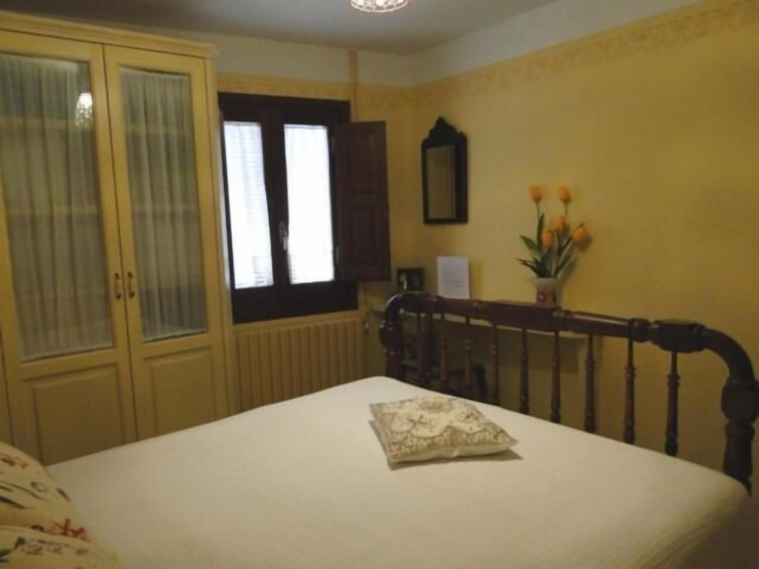 A bedroom with a bed, a wooden headboard, a window with curtains, a radiator, a mirror, and a vase with flowers.