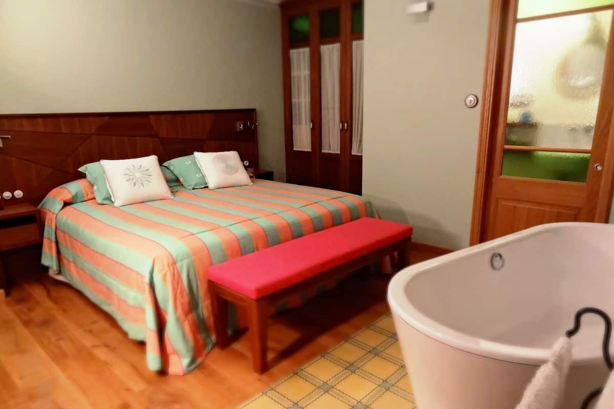 Une chambre à coucher avec une tête de lit en bois, une couvre-lit à rayures, un banc, une baignoire et un sol carrelé.