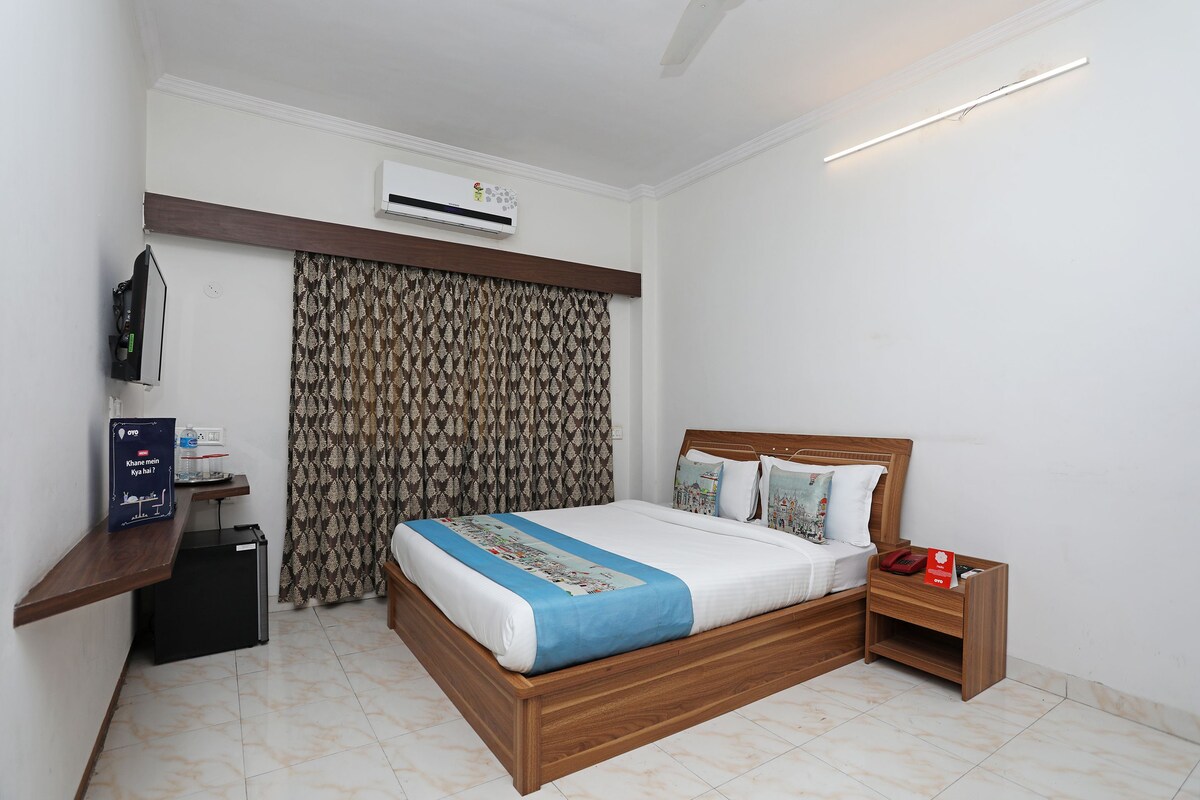 A bedroom with a wooden bed, a nightstand, a desk with a computer, an air conditioner, and patterned curtains.