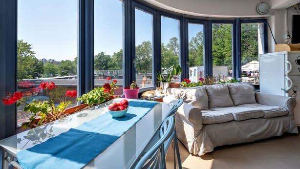 A sunlit room with a balcony, a sofa, a table with a blue tablecloth, and potted plants. - Island Hostel - Adults only (Budapest)