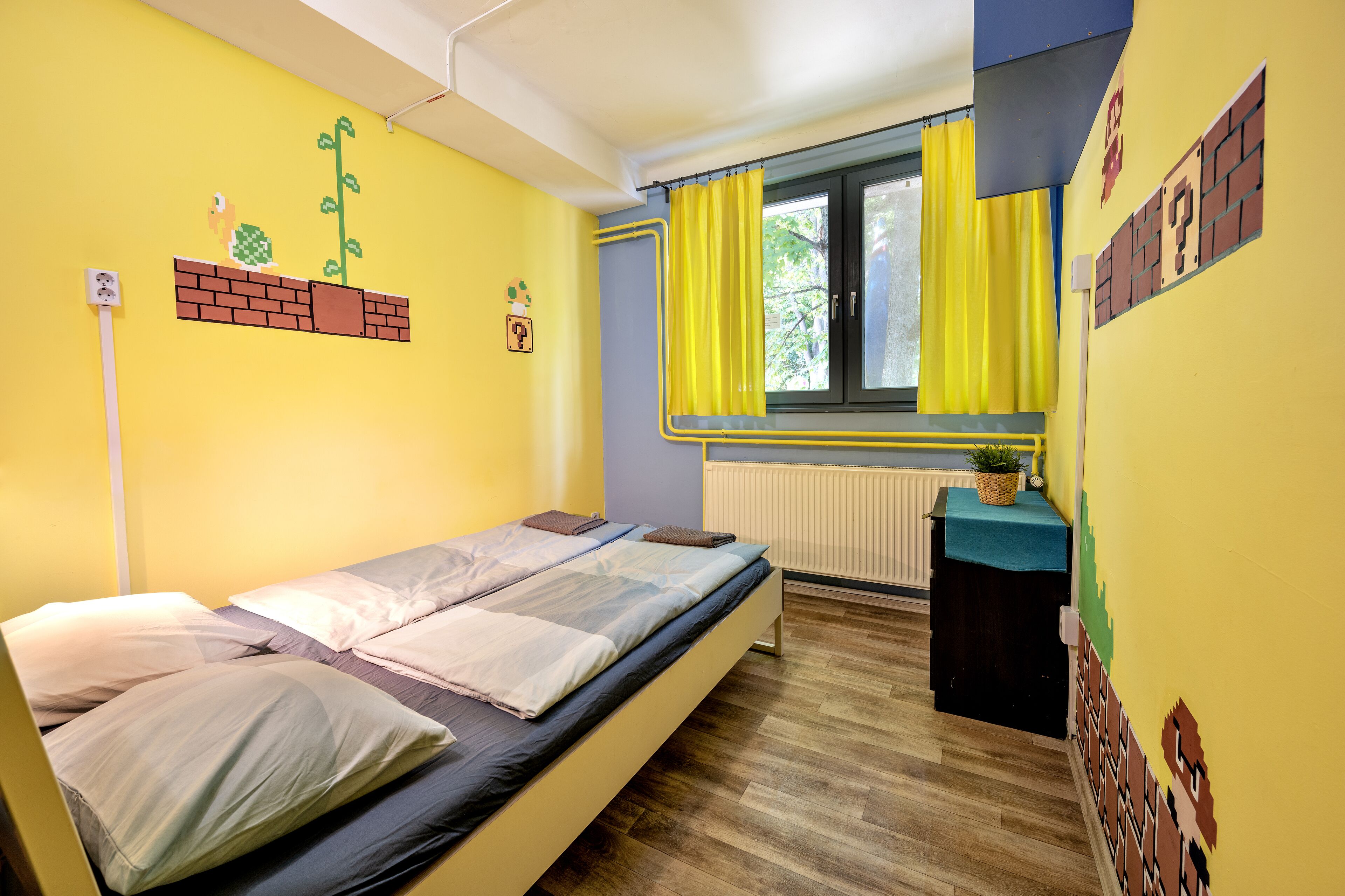 A room with two beds, a window with yellow curtains, a radiator, and a small table with a plant.