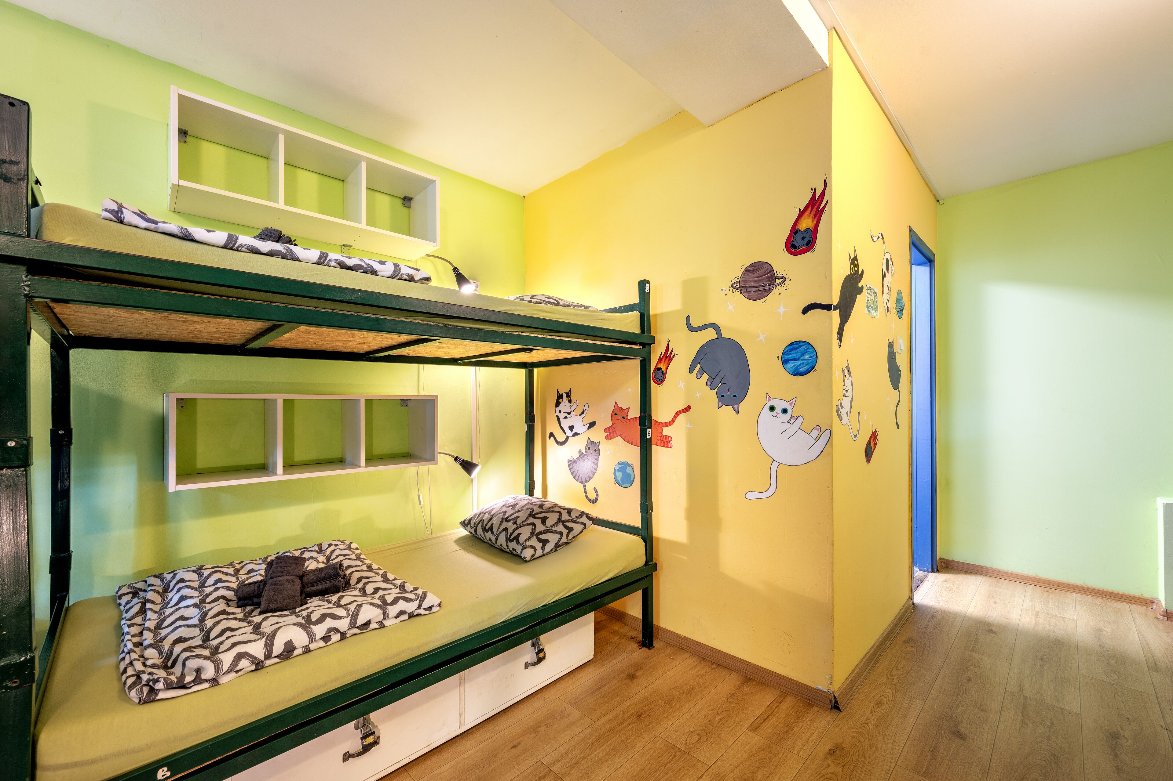 Un lit superposé avec une structure verte et un lit inférieur recouvert d’une couverture à motifs. La pièce a un mur jaune orné d’autocollants représentant des chats de dessins animés.