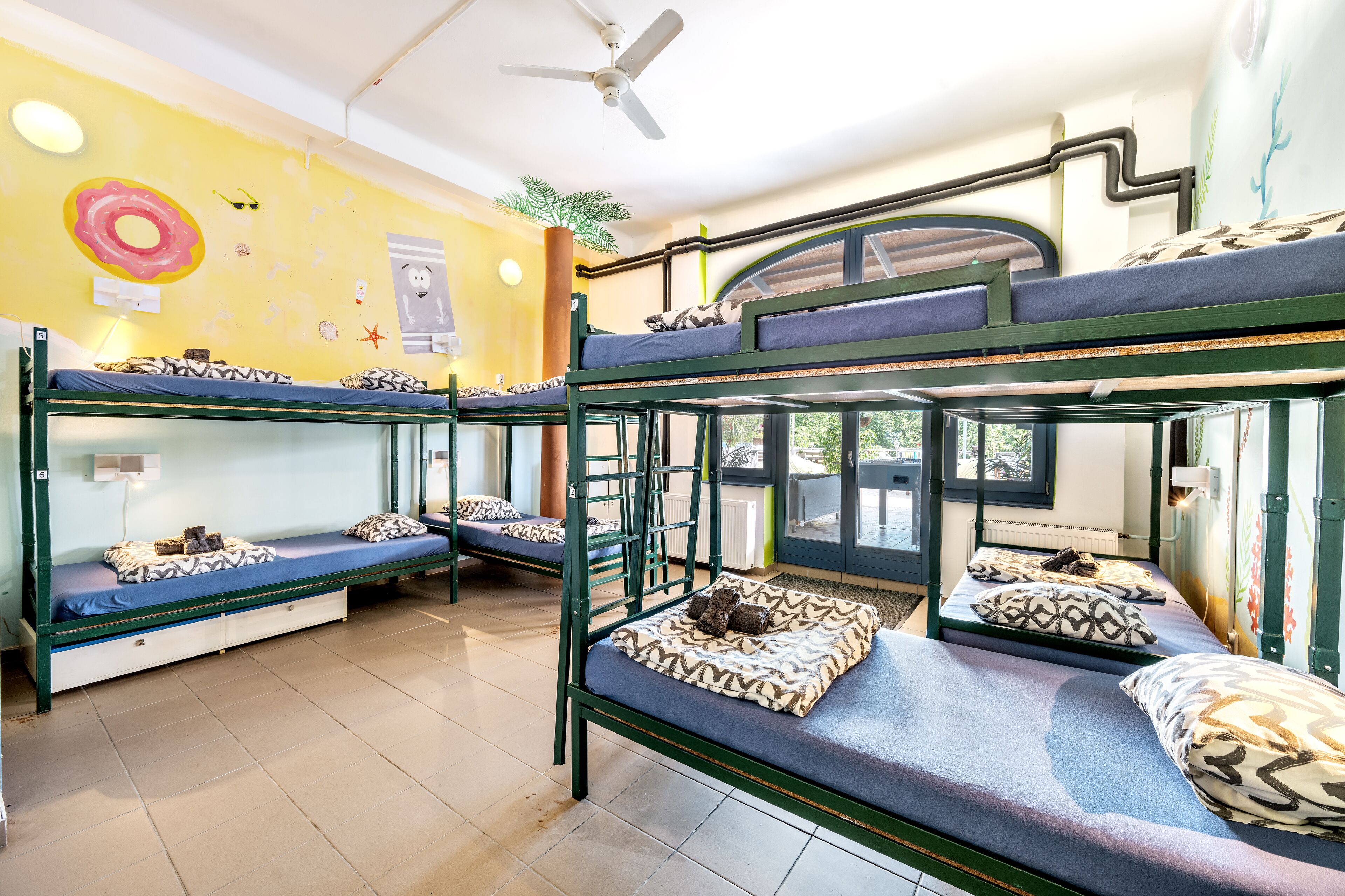 A dormitory room with bunk beds, a ceiling fan, and a window with a view of greenery.