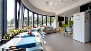 A modern kitchen with a white refrigerator, a dining area with a table and chairs, and a large window with a view of trees. - Island Hostel - Adults only (Budapest)