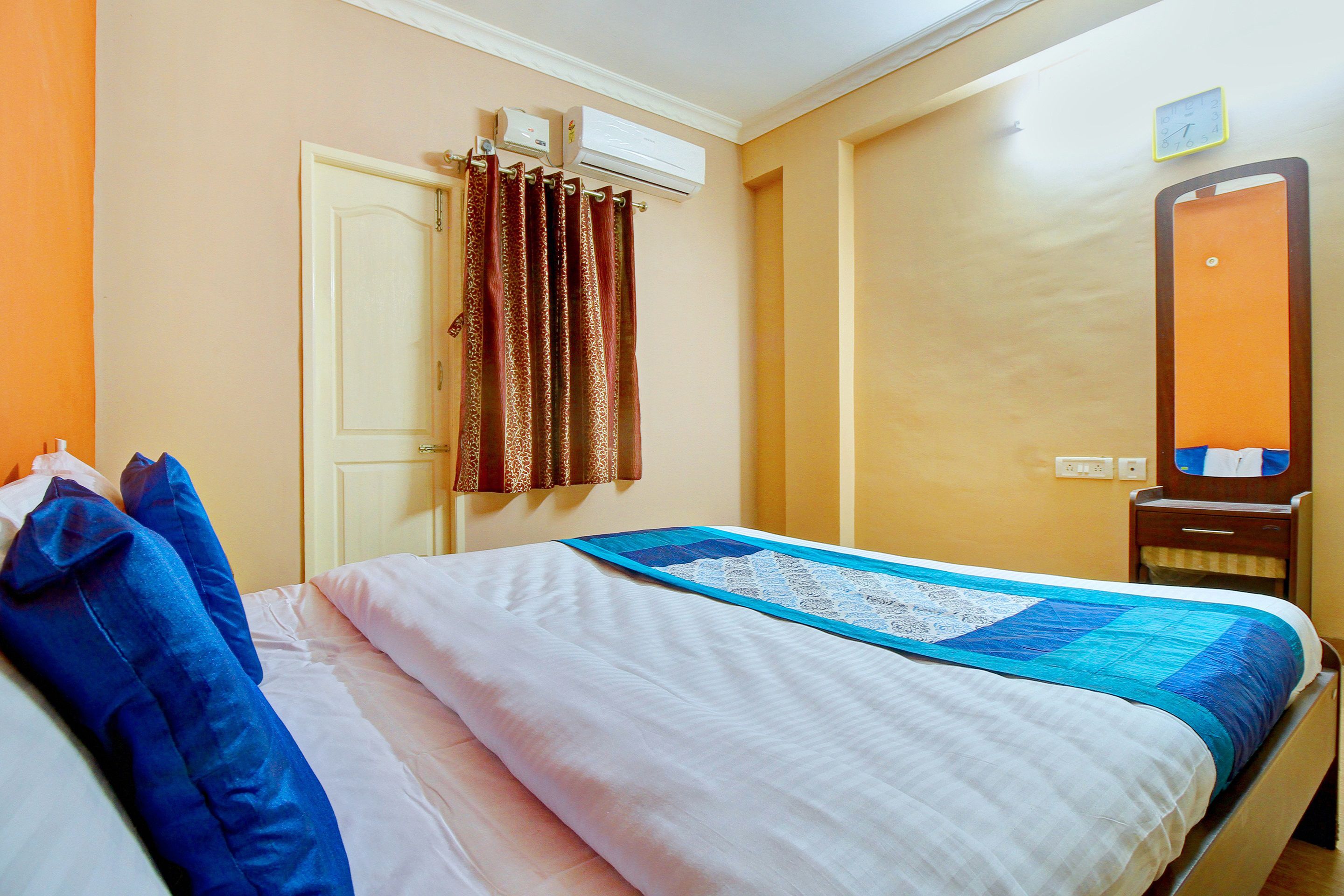 A hotel room with a bed, a nightstand, an air conditioning unit, a mirror, and a door.